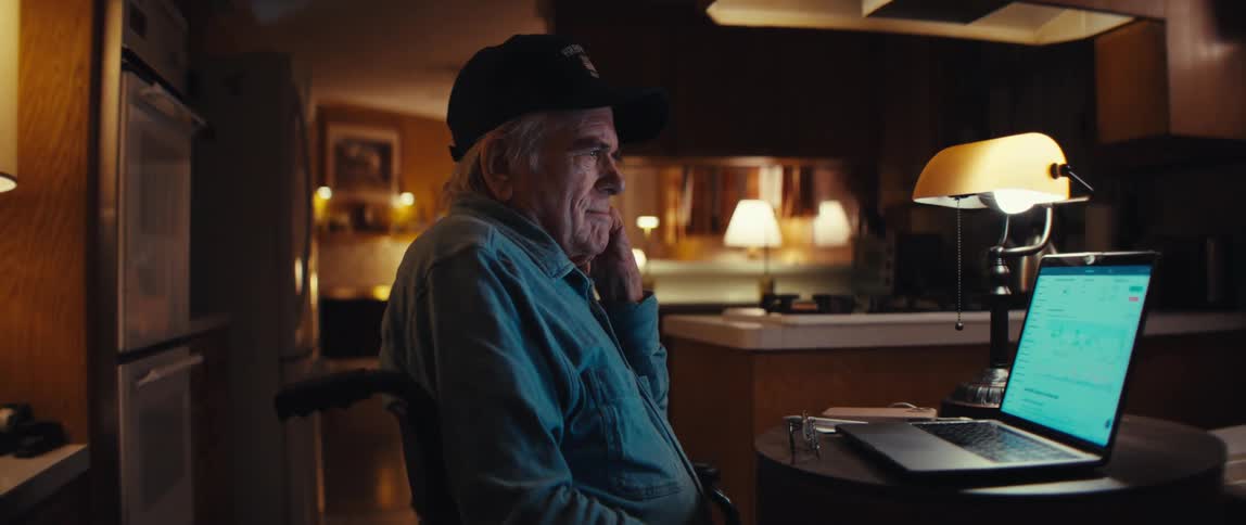 A pensive senior man in a wheelchair sits at his kitchen table at night, thoughtfully watching the glowing screen of his laptop