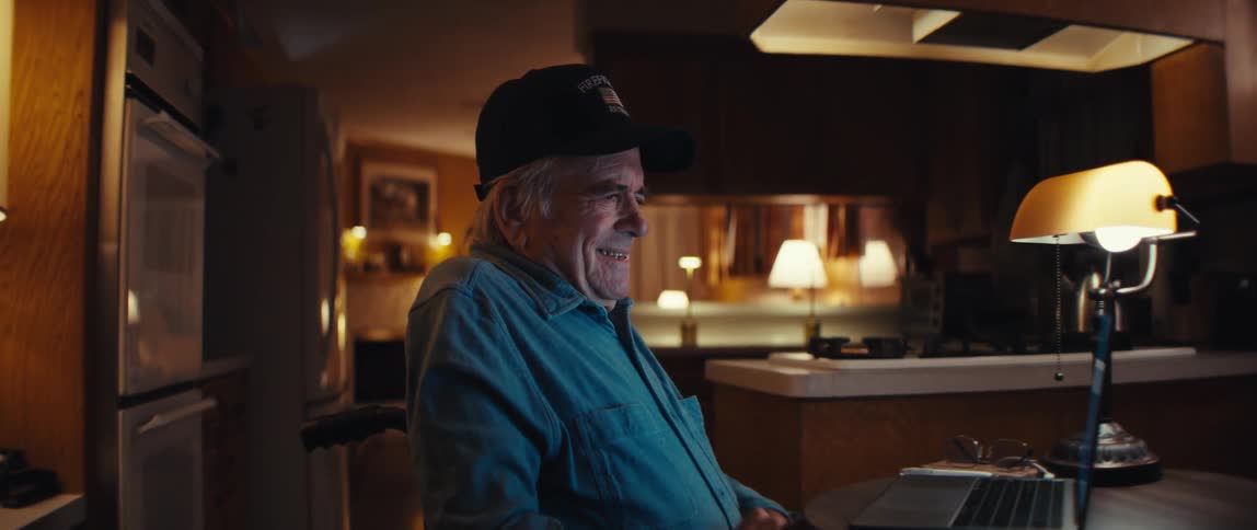 A senior man in a wheelchair happily uses his laptop computer at his kitchen table at night, smiling with satisfaction