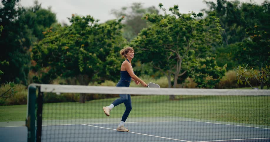 Smiling athletic woman in a blue outfit happily jumps to hit a pickleball with an overhead smash in slow motion