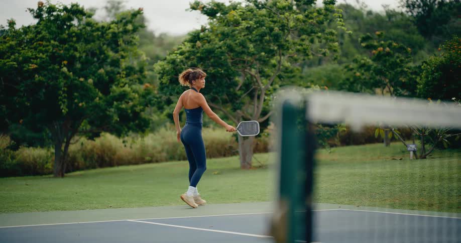 Athletic woman in stylish activewear plays pickleball, hitting a jumping forehand shot in slow motion on a beautiful outdoor court