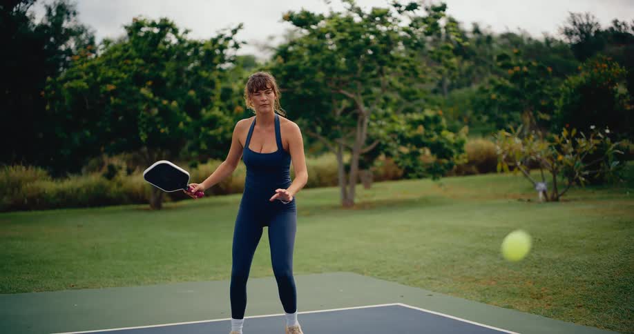 A stylish woman in an athletic jumpsuit practices her pickleball skills in slow motion on an outdoor court surrounded by lush greenery