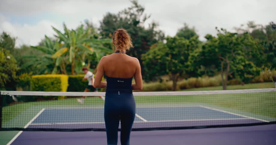 Over-the-shoulder slow-motion view of a woman returning a shot during a pickleball match on a lush, green court