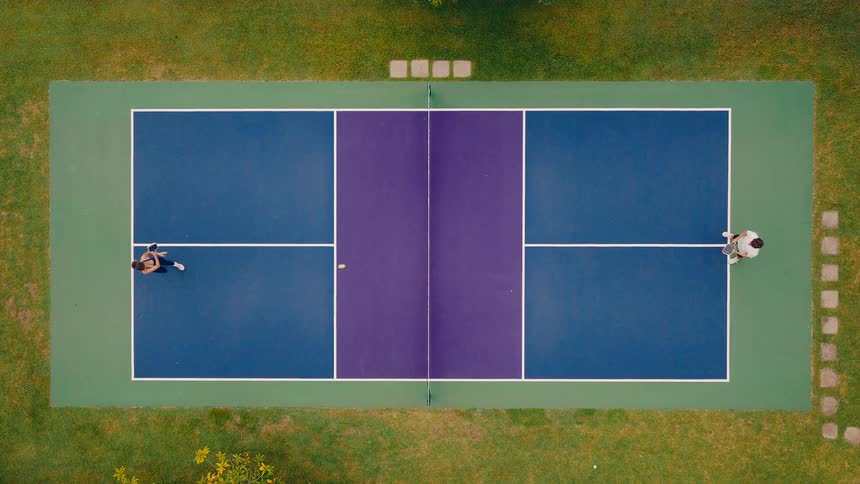 An aerial top-down view shows a man and woman having a slow-motion tennis rally on a colorful court