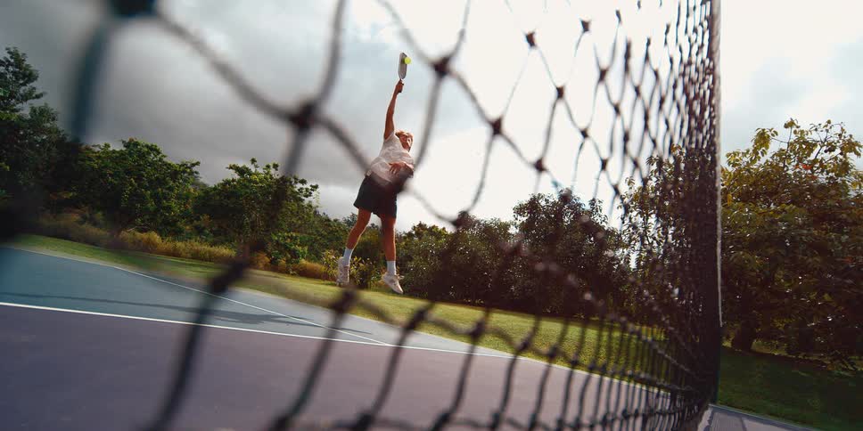 A male pickleball player leaps into the air to hit a powerful overhead shot in slow motion, seen from a low angle through the court net