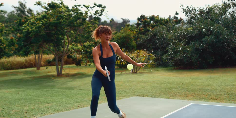Super slow motion of a fit woman in a blue bodysuit hitting a pickleball on a court surrounded by lush greenery