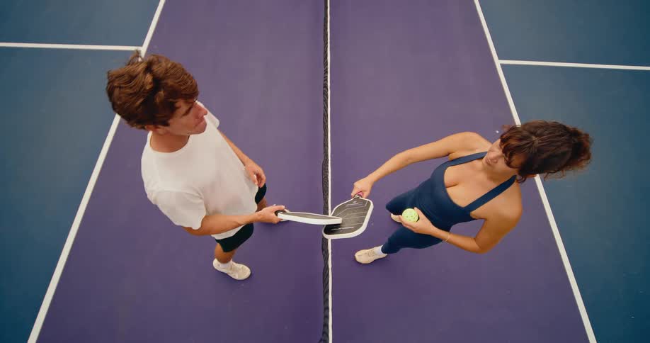 A man and woman face each other across the net on a pickleball court, captured from a slow-motion top-down view