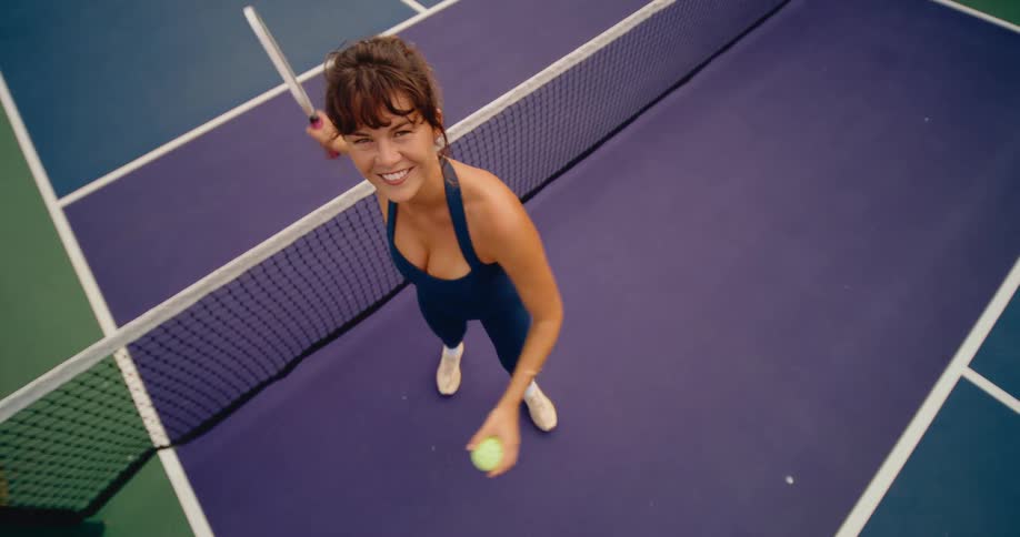 A happy woman in athletic wear tosses a pickleball towards the camera in slow motion on a purple court