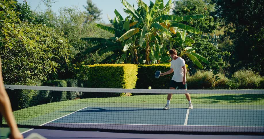 A young man and woman exchange volleys during a friendly game of pickleball on a sunny day in a lush backyard setting