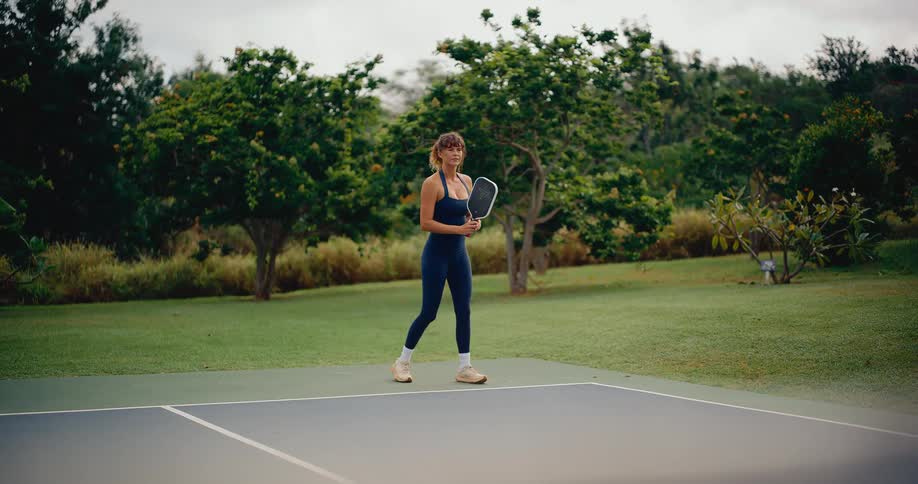 Slow motion wide shot of an athletic woman returning a volley while playing pickleball on an outdoor court surrounded by lush greenery