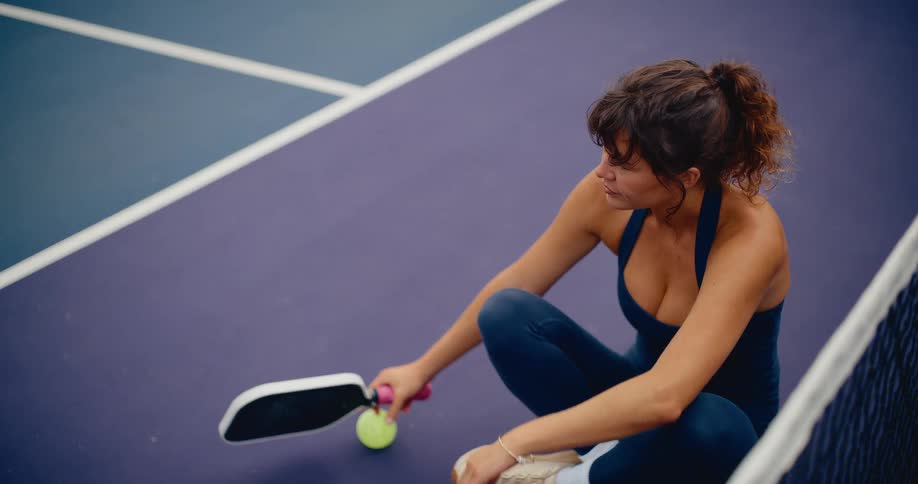 A female athlete relaxes on a pickleball court before picking up her paddle and ball to play a match
