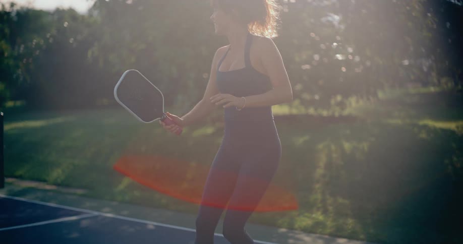 A fit woman in a stylish athletic outfit plays pickleball in slow motion, backlit by the bright sun creating a dreamy lens flare