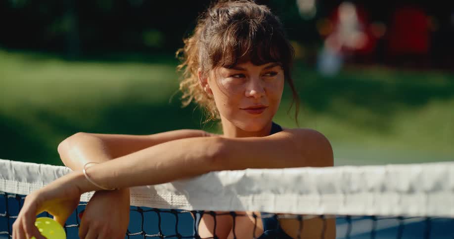 A confident female pickleball player enjoys a moment of rest, leaning against the court net with a ball and paddle in hand