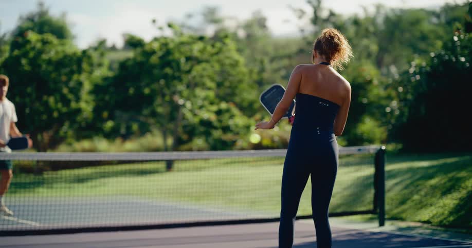 A woman in a black athletic outfit returns a serve while playing pickleball with a man on a beautiful outdoor court in slow motion