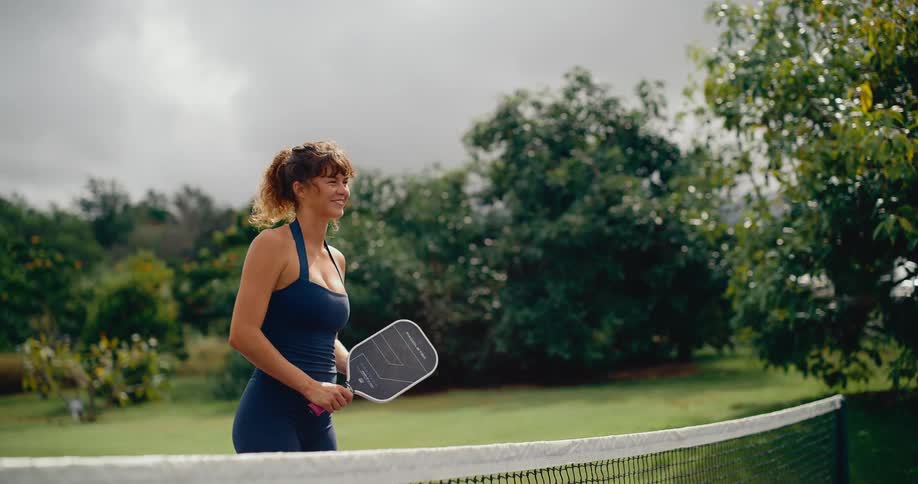 Stylish athletic woman with curly hair smiles as she gracefully bounces a pickleball on her paddle in slow motion on an outdoor court