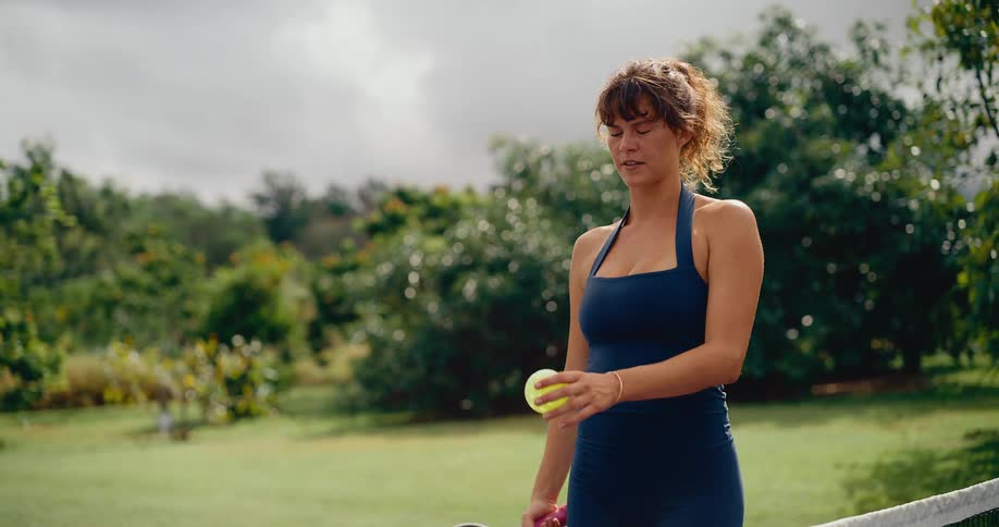 A beautiful woman in a blue athletic outfit stands thoughtfully on a green pickleball court, holding her paddle and ball