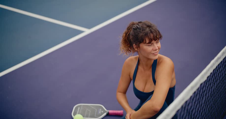 An athletic woman in blue sportswear sits on a purple pickleball court, relaxing and smiling thoughtfully near the net
