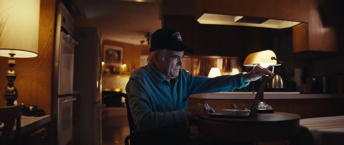 A slow zoom reveals an elderly man in a wheelchair opening a laptop at his table in a dimly lit home, smiling as the screen illuminates his face