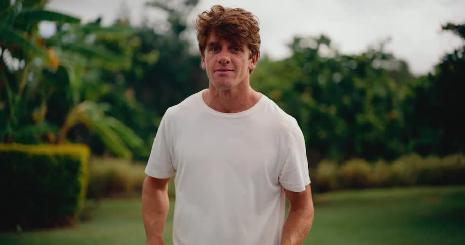 A fit young man in a white t-shirt plays with a pickleball and paddle in slow motion, then looks at the camera with a confident smile