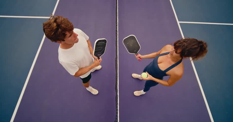 An overhead cinemagraph of a man and woman talking across the net on a purple and blue pickleball court