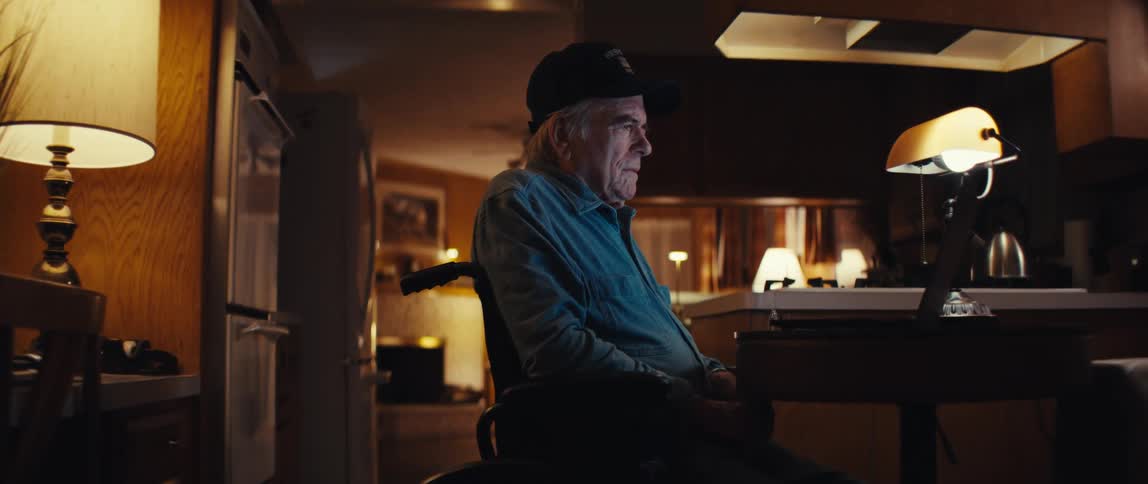 A lonely elderly man in a wheelchair sits in his dimly lit kitchen at night, his face illuminated by the glow of a laptop screen as the camera slowly pushes in