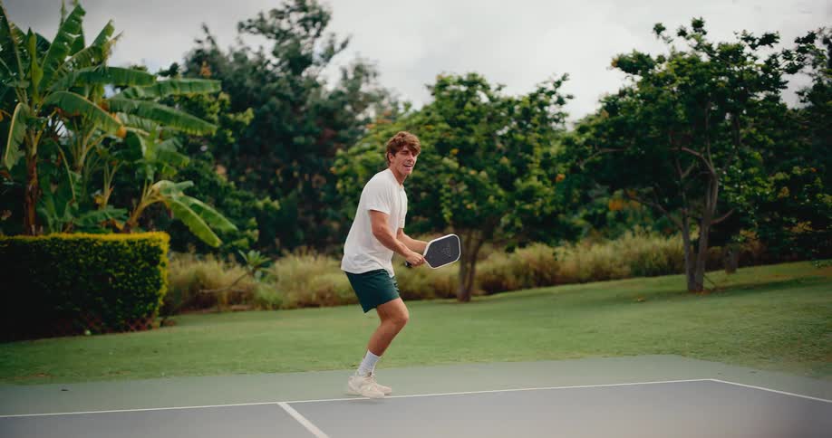 Slow-motion shot of a focused young man playing pickleball on an outdoor court surrounded by lush greenery