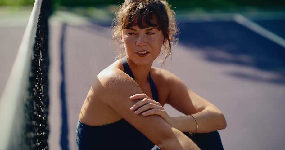 A beautiful female athlete takes a thoughtful break, sitting against the net on a sunny tennis court