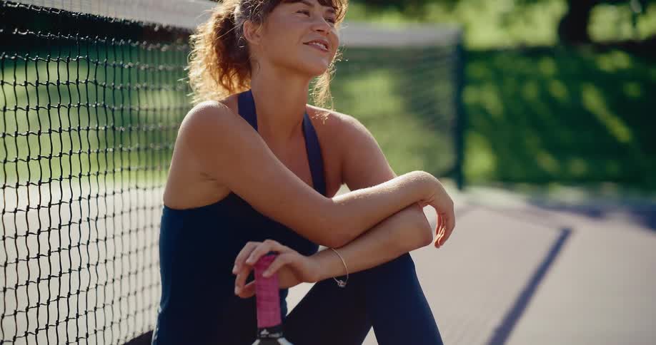 A serene female athlete with curly hair smiles while taking a restful break by the net on a sunny pickleball court