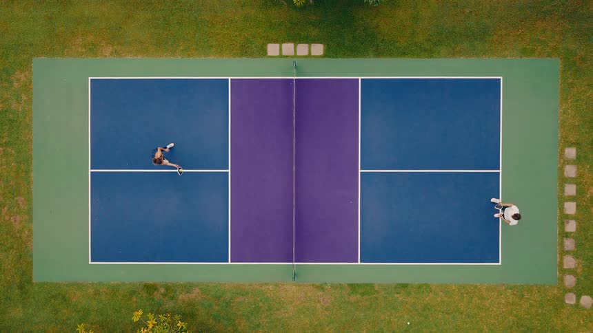 An aerial top-down slow-motion view of a man and woman playing pickleball on a colorful blue and purple court