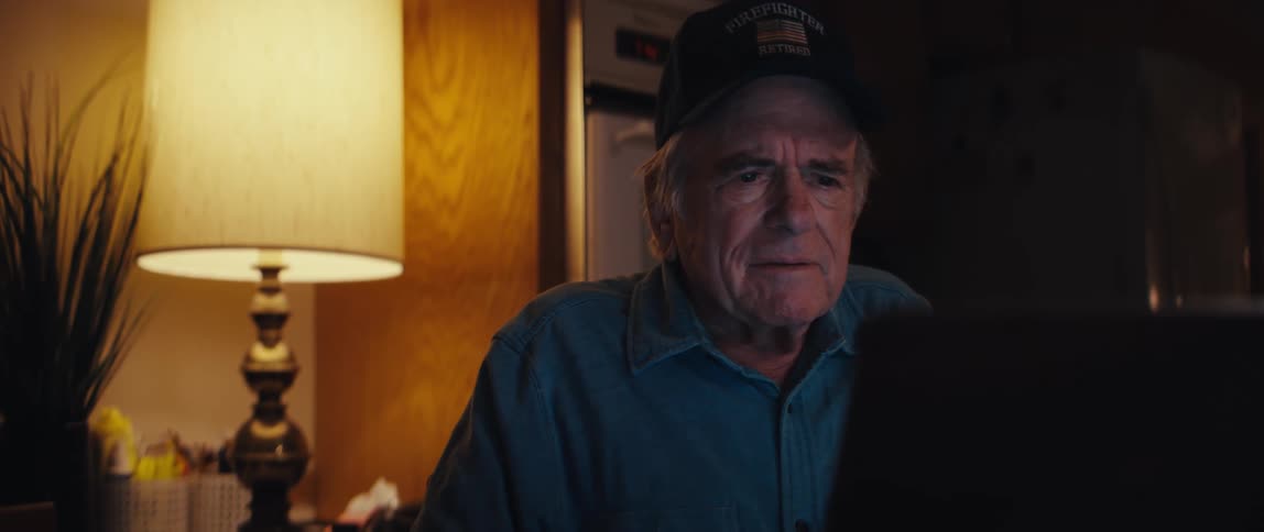 An elderly retired firefighter wearing a cap watches a screen with a sad and reflective expression in his home at night