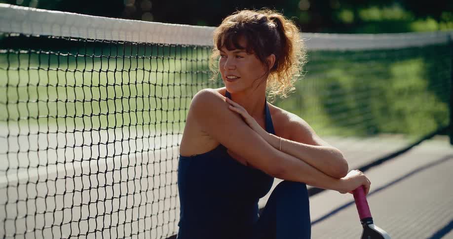A smiling female pickleball player sits on the court and rests against the net on a beautiful sunny day