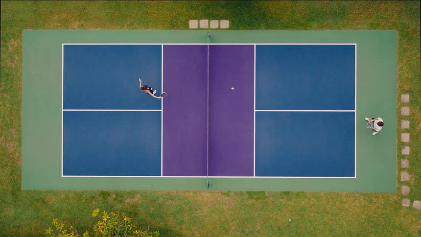An aerial top-down view of a man and woman playing tennis in slow motion on a colorful court