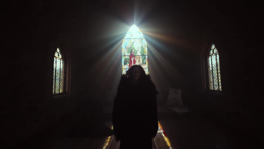 A mysterious cloaked figure slowly walks forward in a dark church, backlit by dramatic sun rays pouring through a stained glass window