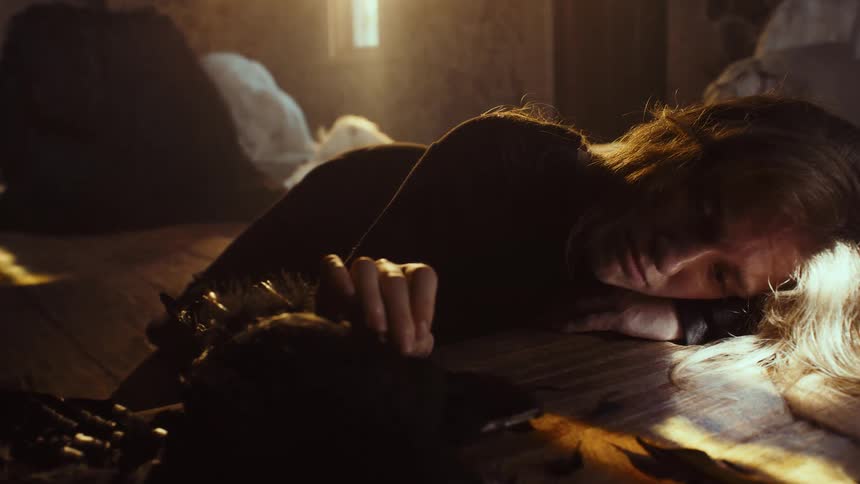 A sorrowful woman lies on a wooden floor, gently touching a deceased black bird in a dramatic beam of light