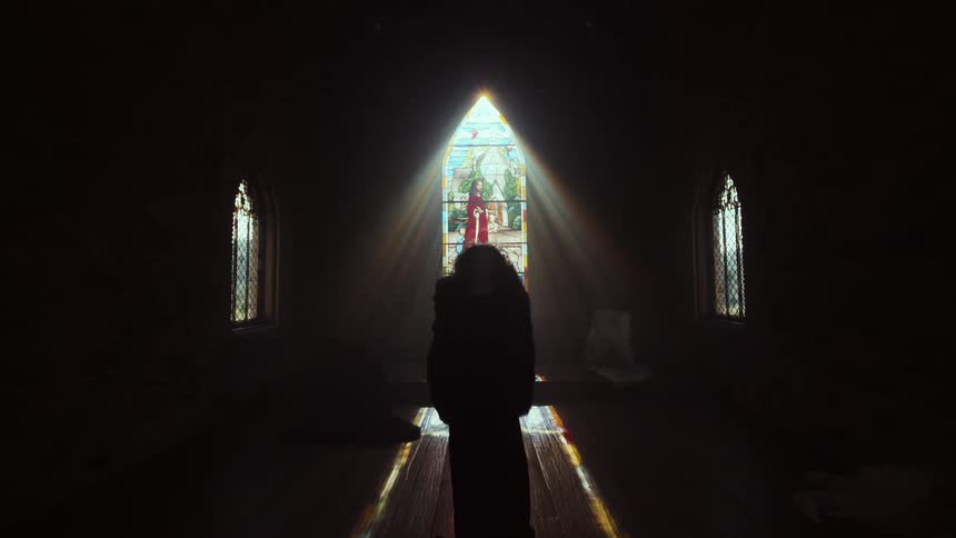 A mysterious cloaked figure is silhouetted by dramatic sun rays streaming through a stained-glass window in a dark church