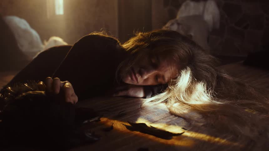A distressed woman with long blonde hair lies unconscious on a wooden floor in a sun-drenched, dusty room next to a broken horned mask