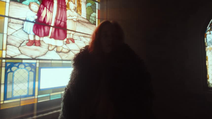 A mysterious woman in a dark fur coat stands in a dimly lit church, gazing at a colorful stained-glass window as beams of sunlight shine through