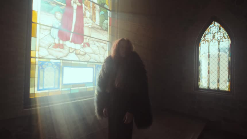 A woman in a dark, furry coat stands in a dimly lit church as dramatic light rays stream through a large stained-glass window