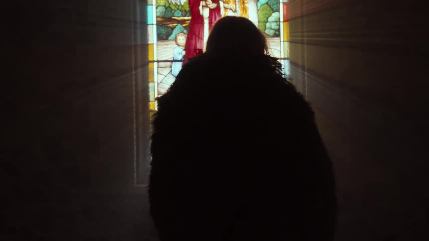 A mysterious silhouetted figure in a furry cloak slowly approaches a beautiful stained-glass window as heavenly sunbeams fill the dark church