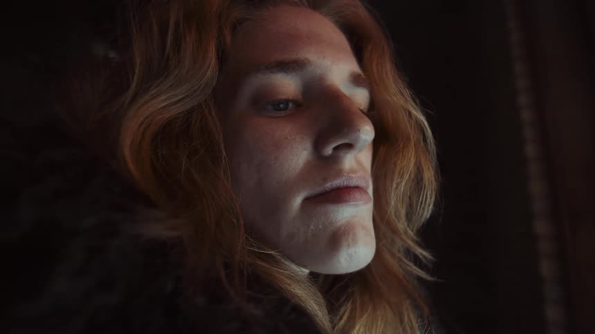 A thoughtful, long-haired person in a heavy coat gazes out of a stained-glass window in a dark, historic church