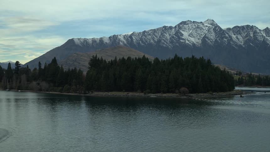 An aerial drone shot flies over Lake Wakatipu in Queenstown, New Zealand, where a jet boat speeds past a forested island with the snow-capped Remarkables mountains in the background