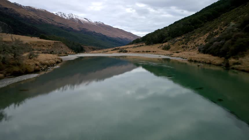 Aerial drone footage flies down a beautiful turquoise river winding through a valley with snow-capped mountains in the distance