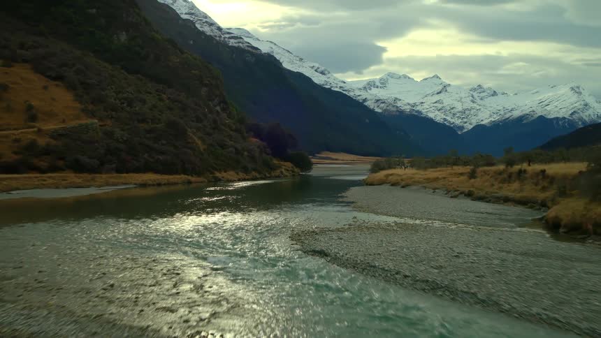 An epic aerial flight down a wide river valley towards stunning snow-capped mountains