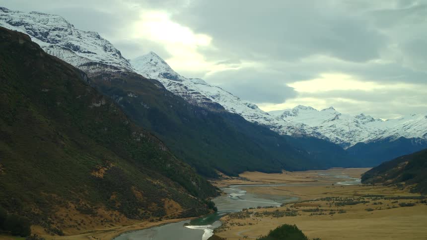 An epic aerial shot reveals a vast mountain valley with a winding river and majestic snow-capped peaks under a dramatic cloudy sky