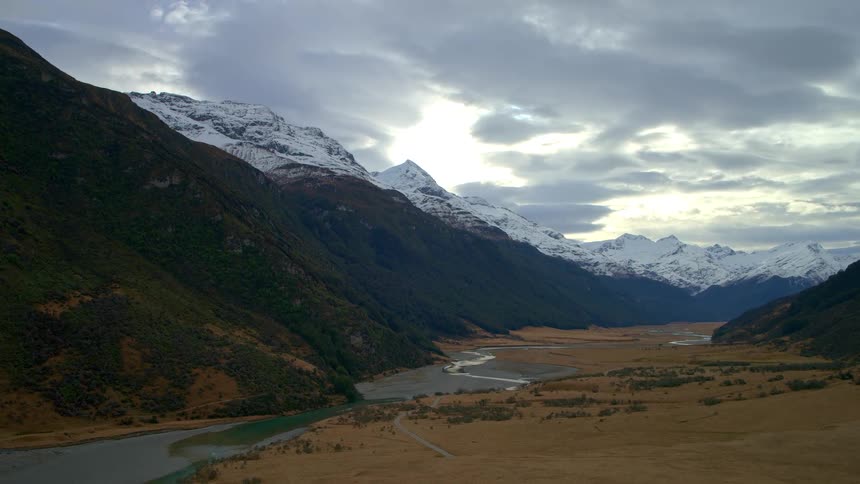 A slow aerial flyover of a dramatic valley floor with a winding river leading to towering snow-capped mountains