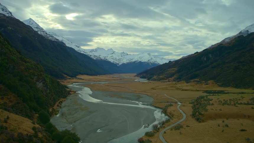 An aerial view tilts up from a rushing river to reveal a vast valley surrounded by snow-capped mountains