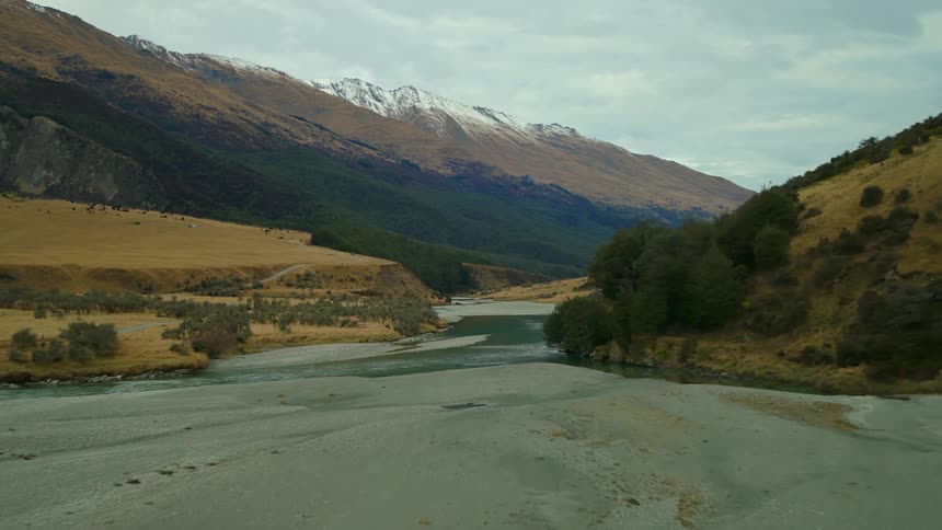 An aerial drone shot flies forward over a wide, braided river flowing through a vast mountain valley with snow-capped peaks in the distance