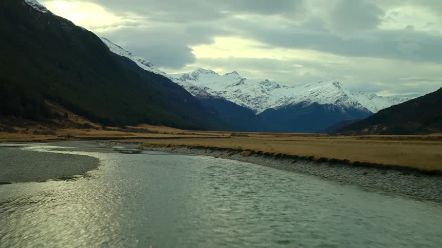An epic aerial flight follows a winding river through a vast valley towards a majestic range of snow-capped mountains