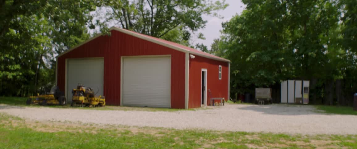 A red metal barn with two large garage doors sits on a gravel driveway in a rural setting with trees