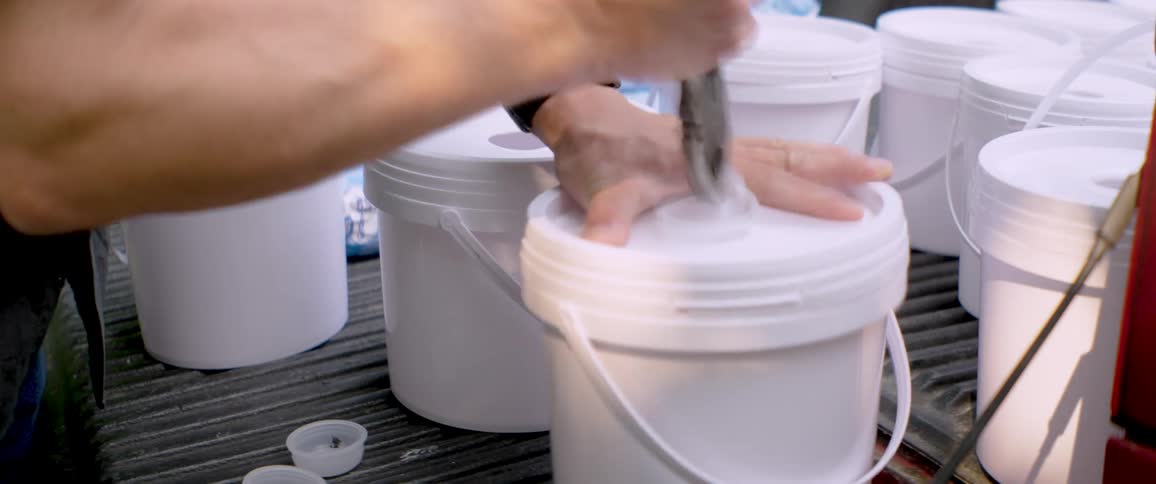 A beekeeper prepares bee feeder buckets by punching holes in the lids with pliers on the tailgate of a truck