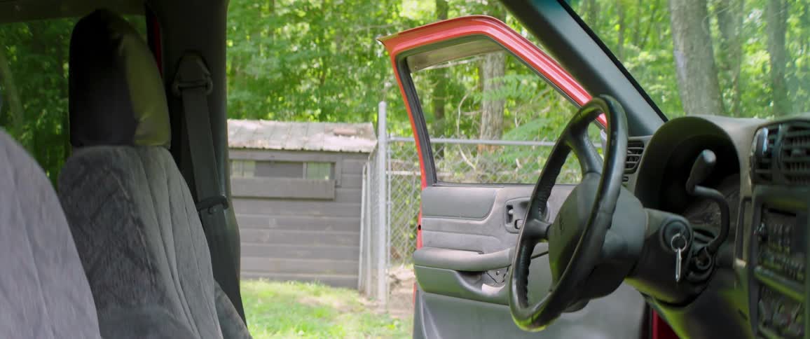 The view from inside a red pickup truck with the passenger door ajar, revealing a rural backyard with a shed and dense green trees